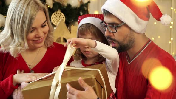 Father and Mother in Red Santa Hats Give Gift to Child Girl