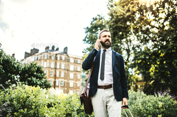 Hipster businessman with smartphone and suitcase walking in park in London. - Stock Photo - Images