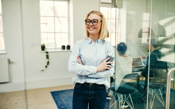 Smiling businesswoman leaning with her cellphone against an office wall - Stock Photo - Images