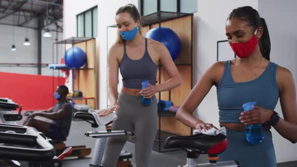 Two diverse women disinfecting exercise bike saddles at gym alt