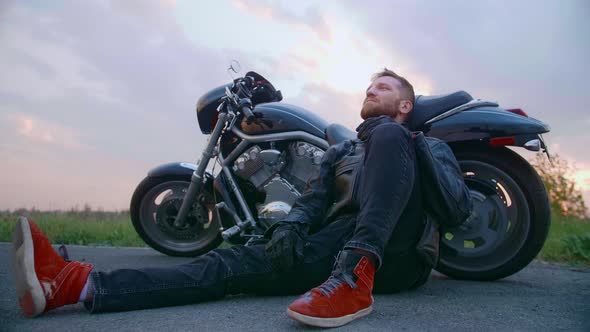 Middleaged Biker with Beard Sits By Motorcycle at Sunset