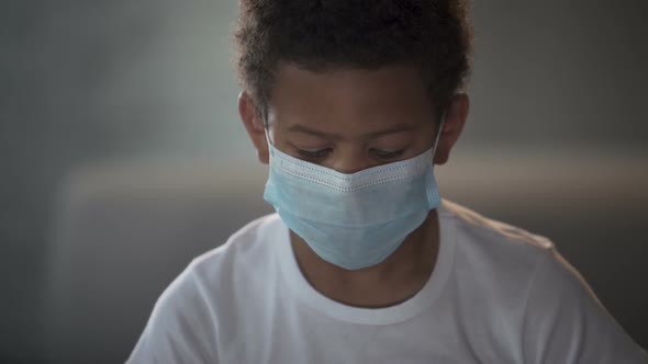 African American Child in Medical Mask Looking Into Camera With Sad Eyes alt