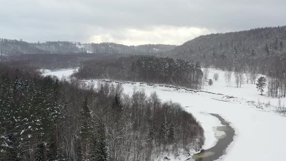 Aerial Drone flight over snowy frozen river in mountain forest valley of Sigulda, Latvia