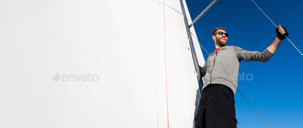 Yacht captain with a beard stands on sail boom on a sailing yacht, holding the rope in his hand and - Stock Photo - Images