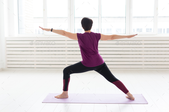 People, sport and healthcare concept - Middle-aged woman practicing yoga, standing in Warrior two