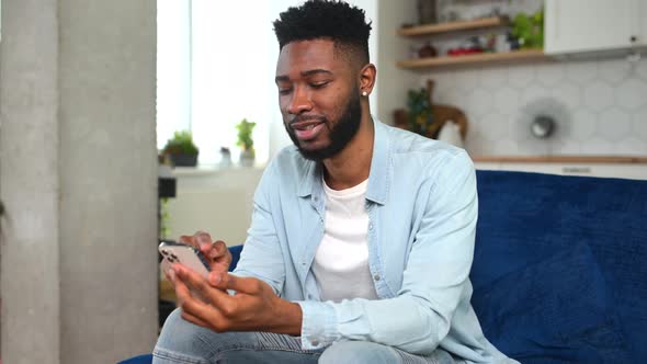 Cheerful Young Black Guy Sending Message on Mobile Phone While Sitting at the Sofa alt
