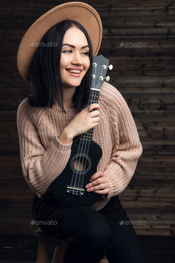 Young smiling with a ukulele sit in studio on a wooden background Stock by romankosolapov