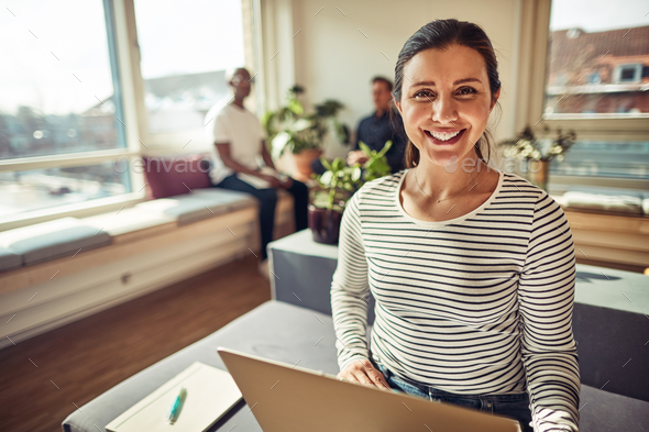 Smiling businesswoman using a laptop with coworkers in the background - Stock Photo - Images