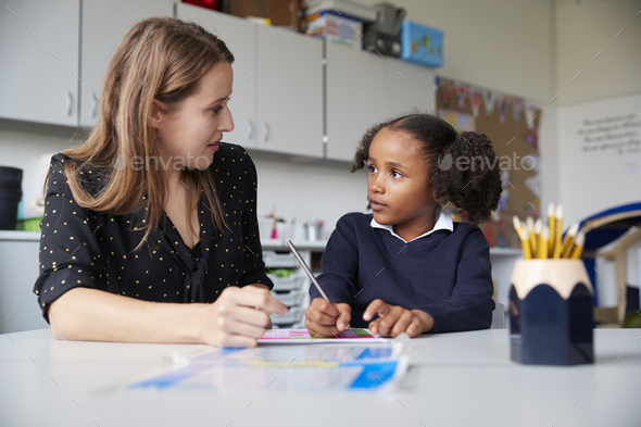 Young female primary school teacher working one on one with a schoolgirl at a table in a classroom - Stock Photo - Images