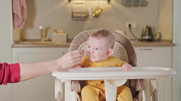 Mother feeding toddler baby with spoon on high chair for children, home kitchen background