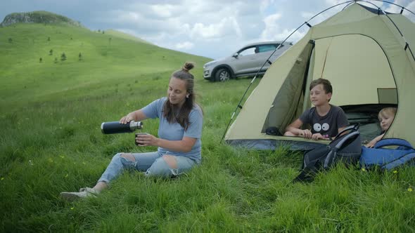 Happy millennial woman in blue t-shirt pouring hot tea from thermos against tent with kids.