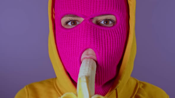 Close up of young woman in pink balaclava and yellow hood eats banana on purple background. alt