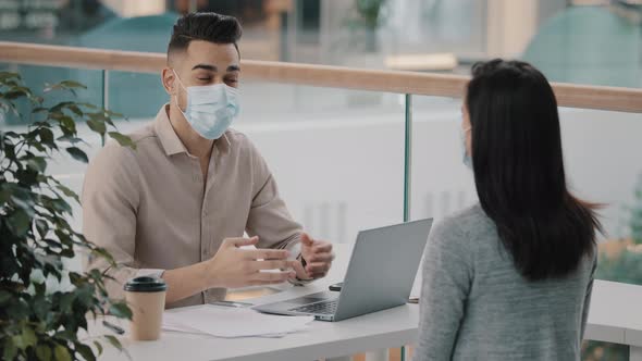 Young Hispanic Bank Worker in Medical Face Mask Telling Unrecognizable Client Woman Benefits Using alt