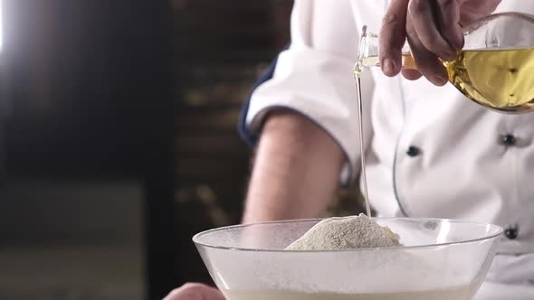 chef in kitchen pours olive oil from a bottle in a thin stream into wheat flour. alt