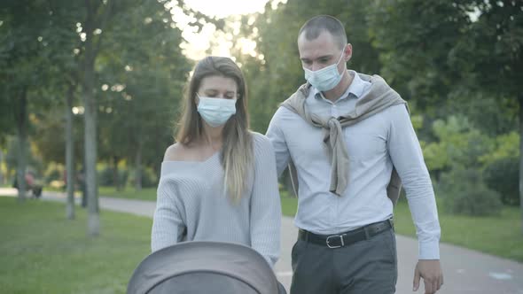 Young Caucasian Man and Woman in Covid Face Masks Strolling at Sunset with Baby Carriage and Talking alt