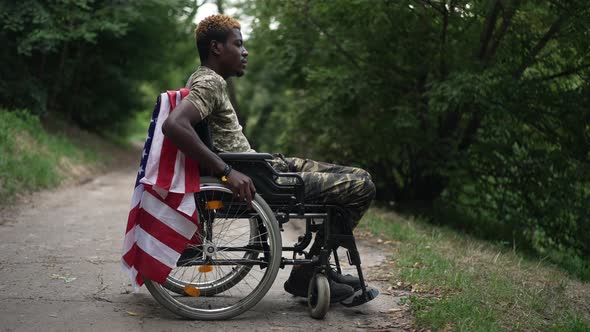 Side View Thoughtful Depressed African American Young Soldier in Wheelchair with USA Flag Looking alt