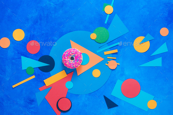 A pink donut with colorful paper decorations. Color blocking flat lay with sweets. Complementary