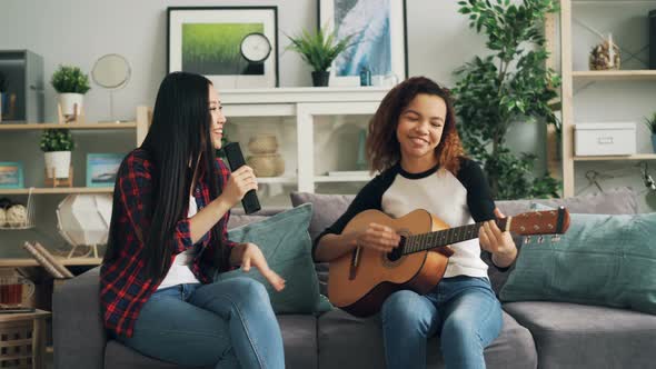 Female Friends Are Having Fun at Home Playing the Guitar and Singing in TV Remote Control Enjoying alt