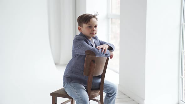 A Teenage Boy in a Jacket and Jeans Poses on a Chair By a Window alt