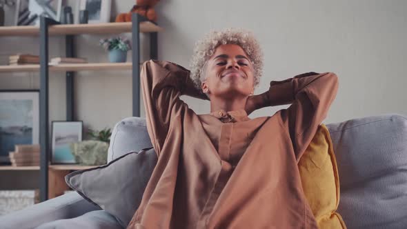 African Woman Resting on Couch Taking Deep Breath Holding Hands Behind Head alt