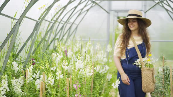 Woman in Straw Hat Smells Flowers Walking Along Greenhouse
