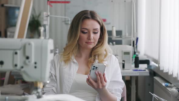 Young Beautiful Light Hair Seamstress Uses Smartphone in the Workplace
