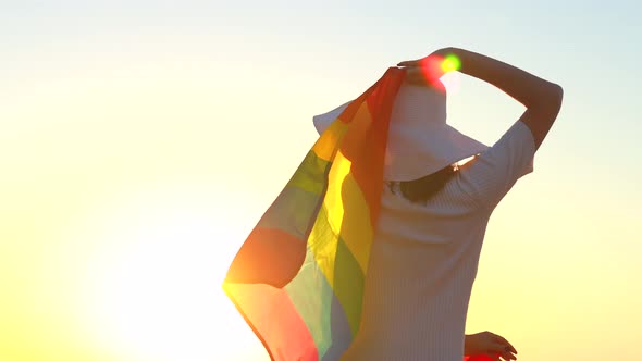 Back View of Woman in White Dress and Hat Holding Gay Pride Flag at Sunset in Beach alt