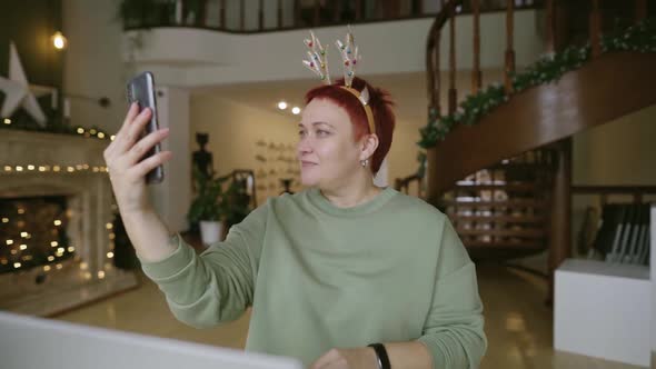 Adult Woman Sitting at Workplace at Home and Taking Selfie with Christmas Horns alt