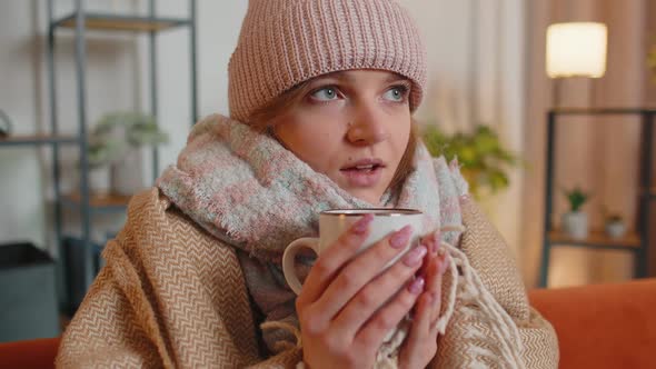 Young Sick Woman Wear Hat Wrapped in Plaid Sit Alone Shivering From Cold on Sofa Drinking Hot Tea alt