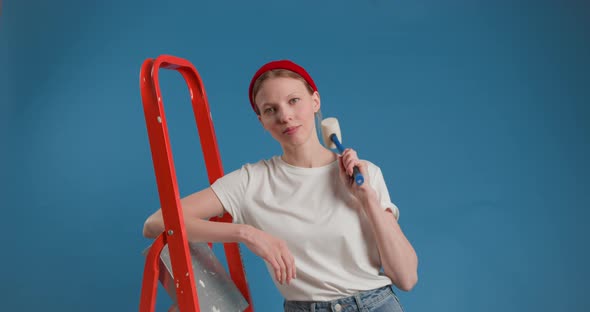 Young Funny Woman in Red Cap Stands with Stepladder Holding Hammer in Hand on Blue Background alt