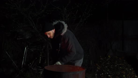 Homeless Young Man Stands By the Fire. A Man Stands at Night Near a Barrel of Fire and Bask. alt