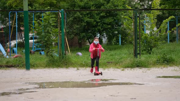 A little girl in a medical mask rides a scooter through puddles on a spring day after rain.