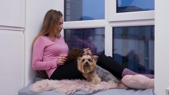 Young Woman Sits on Windowsill Near Large Windowenjoys Reading an Interesting Book alt