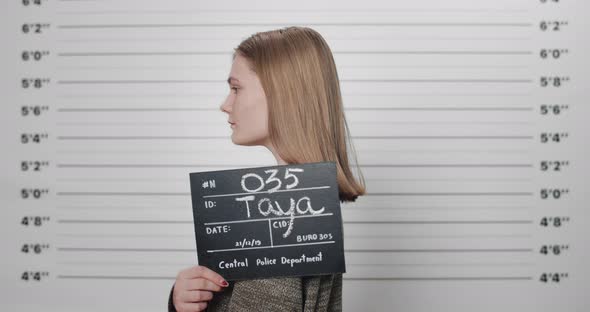 Mugshot of Arrested Female Person Holding Sign While Being Photographed in Front of Metric Lineup alt