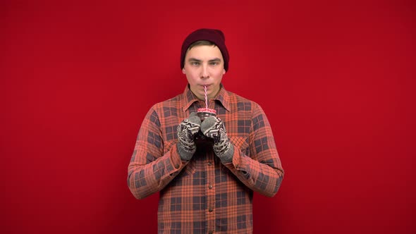 A Young Man in a Hat and Mittens Drinks Mulled Wine. Shooting in the Studio on a Red Background. alt