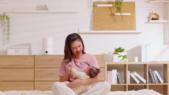 Beautiful Asian mom woman holding newborn baby in her arms sit on bed at white cozy home alt