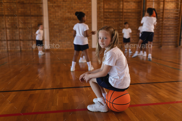 Side view of schoolgirl sitting on basketball and looking at camera at basketball court in school