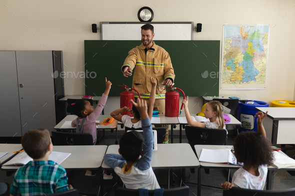 Schoolkids raising hands while firefighter teaching about fire safety of elementary school - Stock Photo - Images