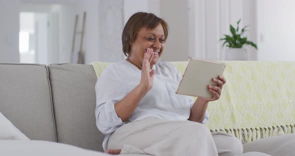 Happy african american senior woman sitting on couch making video call using tablet, waving alt