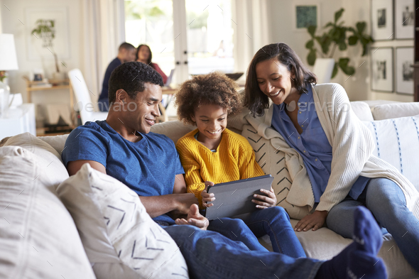 Young parents and their pre-teen daughter sitting on a sofa in the living room using tablet computer - Stock Photo - Images