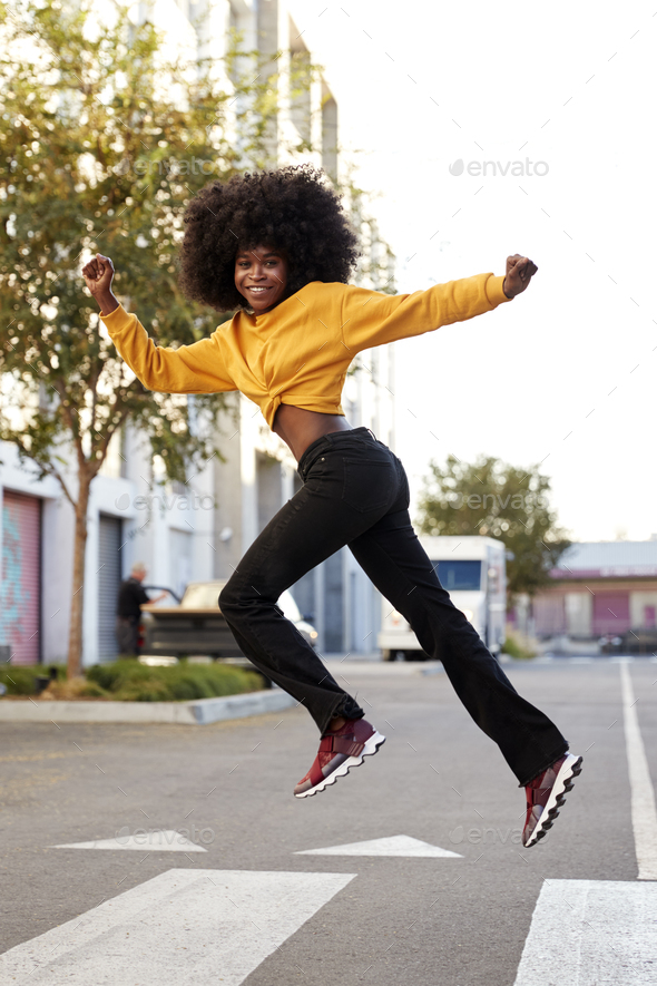 Young black woman with afro jumps, looking to camera, while crossing street, side view, vertical
