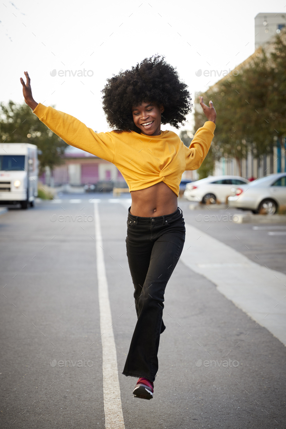 Young black woman with afro jumps, looking to camera, while crossing street, front view, vertical