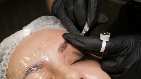Beautician in Black Gloves Does Permanent Makeup Correction of the Shape of the Eyebrows to Young alt