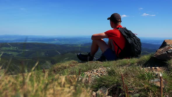 Young Man Sits on the Edge of the Mountain and Looks Around - Valley with Trees and Blue Sky alt