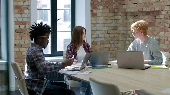 Wide Shot of Young Caucasian and African American Colleagues Sitting with Laptops in Office and alt