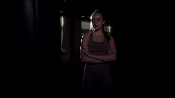 Young Confident Woman with Crossed Hands Standing in Darkness at Punching Bag Smiling Looking Away alt