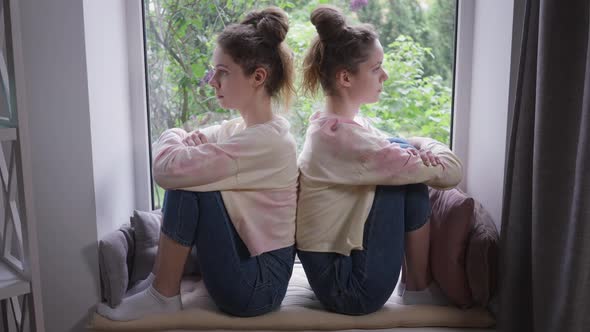 Side View Two Identical Young Women Sitting on Windowsill Back to Back Thinking alt