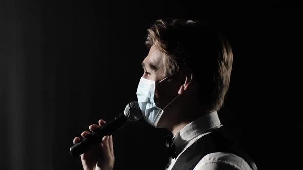 Man Host of Event in Medical Mask Speaks at Microphone at Concert on Stage.