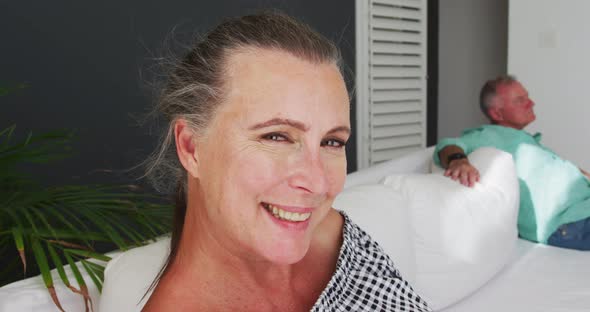 Caucasian senior couple sitting on sofa in the sun the woman smiling and man in background alt