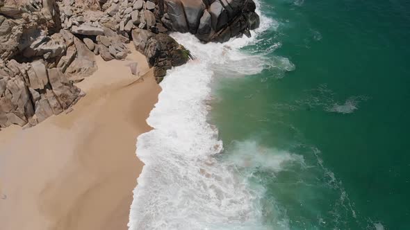 Aerial View of Waves Crashing onto Beach Alongside Rock Formation alt